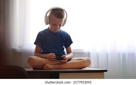 Child's dependence on a mobile device theme. Child with headphones on neck is sitting on table and uncontrollably playing online games on smartphone. virtual life. children's addiction to online games - Shutterstock ID 2177952055