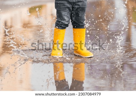 Child's feet in yellow rubber boots jumping over a puddle in the rain.