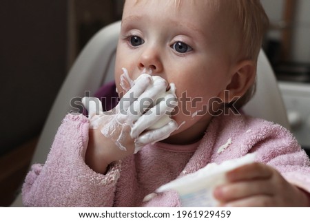 The child's first attempts to eat on their own. The baby girl licks her hands soiled with curd. Grimy child. Yogurt for lunch