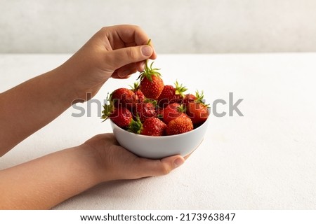 A child's hand holding strawberries on a light background, a plate with strawberries. The concept of summer healthy eating. Side view