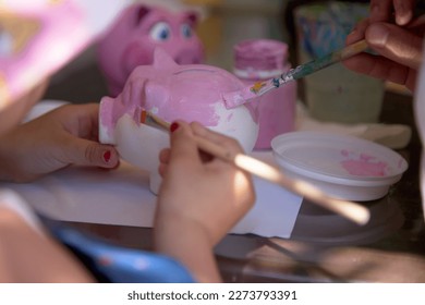 Child's hand holds brush   paints clay piggy bank in the shape pig