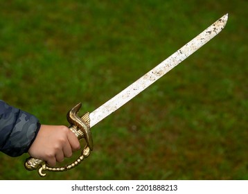 A Child's Hand Holds A Pirate Saber. A Child's Hand With A Pirate's Golden Saber. Sword Fighting.