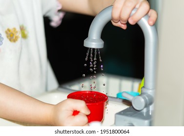 A child's hand holds a red plastic children's toy mug and draws water from the tap into it in close-up. The child plays at home in the kitchen. A toy set for washing dishes. Happy childhood
