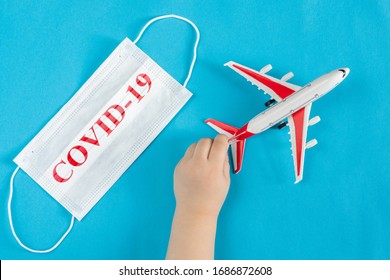 A Child's Hand Holds A Toy Airplane, Next To A Medical Mask With The Inscription Covid-19. Evacuation Of People From Areas Infected With Coronavirus.