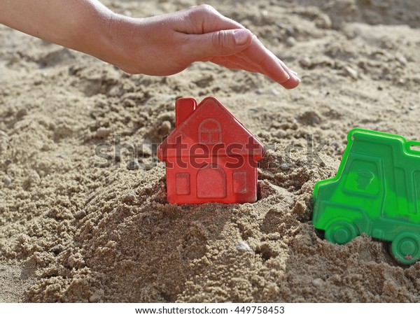 Child\'s hand over the house in a sandbox. Hand\
as a roof over the house. Bright plastic toys in the sandbox or on\
the beach. Sandbox, children, hand, home, house, buy, purchase,\
housing, business.