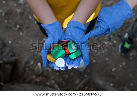 Child's hands with gloves full of plastic caps. Hands of young boy holding a bunch of bottle caps ready to recycle and be ecologically friendly. Another child's hand pointing.