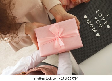 Child's Hands Hold Beautiful Pink Gift Box With Ribbon. Pink Tulip Flower And Sign I Love You Mom In Background. Top View, Close-up. Preparing For The Holidays.