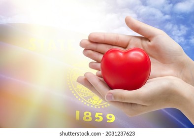 Child's Hands Hold A Heart On The Background Of The Flag Of State Of Oregon, Health Care Concept.