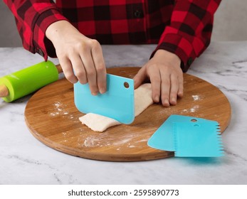Child's hands using blue scraper to cut dough into pieces on wooden cutting board surrounded by rolling pin and other scrapers