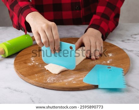 Child's hands using blue scraper to cut dough into pieces on wooden cutting board surrounded by rolling pin and other scrapers