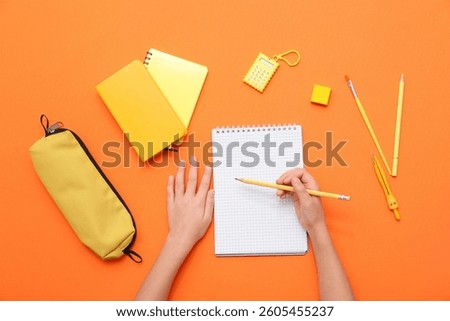 Child's hands writing down something in notebook on orange background. Top view