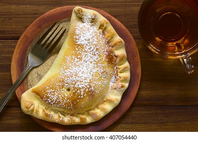 Chilean Apple Empanada With Icing Sugar, Cup Of Tea On The Side, Photographed Overhead On Dark Wood With Natural Light