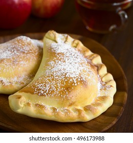 Chilean Apple Empanada With Icing Sugar, Cup Of Tea In The Back, Photographed On Dark Wood With Natural Light (Selective Focus, Focus One Third Into The Image)