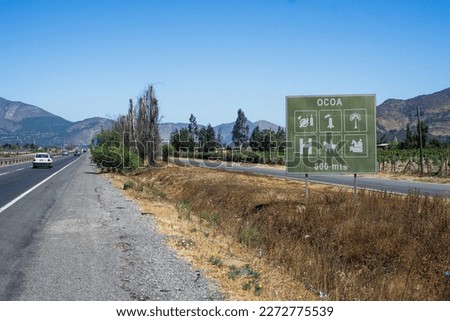 Chile's Route 5 showcases a picturesque view of the surrounding hills and blue sky. A traffic sign in the foreground points 800m towards the rural town of Ocoa, displaying useful tourist information.