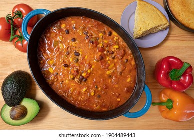 Chili In A Cast Iron Pot With Corn Bread, Avocado, Tomato, And Bell Peppers On A Wood Countertop