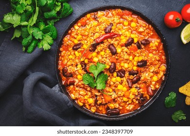 Chili con carne with beef, red beans, paprika, sweet corn and hot peppers in tomato sauce, spicy tex-mex dish in frying pan, black table background, top view