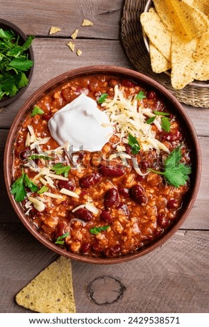 Chili con carne with cheese topings and nachos on wooden table, top view. Homemade mexican food, chili with red beans and ground beef. Chilean beans.
