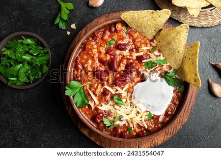 Chili con carne with cheese topings and nachos on black background, top view. Traditional mexican food, chili with red beans and ground beef. Chilean beans.