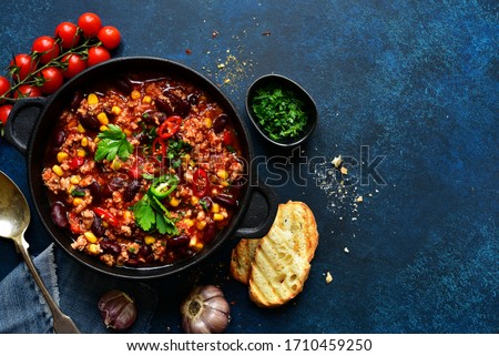 Chili con carne - traditional mexican minced meat and vegetables stew in tomato sauce in a cast iron pan on a dark blue slate, stone or concrete background. Top view with copy space.