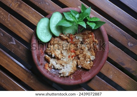 Chili shrimp paste and fried tempeh penyet with two slices of cucumber and basil leaves in a clay mortar container, photo taken at high angle with a lacquered wooden background      