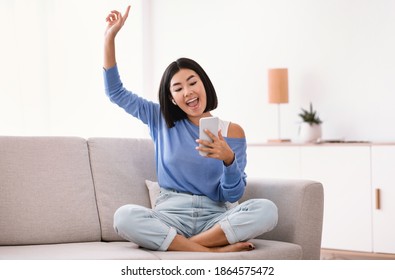 Chill And Lifestyle Concept. Exited asian woman wearing wireless earphones listening to favorite song, holding mobile phone, sitting on sofa and dancing in living room, raising hand and finger up