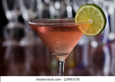 chilled cosmopolitan cocktail served in a  martini glass on a busy bar with a shallow depth of field with blurred glasses and lights in the back, garnished with a lime - Powered by Shutterstock