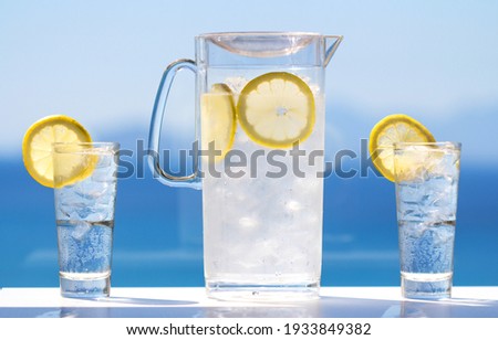 A chilled glass jug and two chilled glasses filled with cold water, ice cubes and lemon slices under bright sun light in front of a blurry sea view
