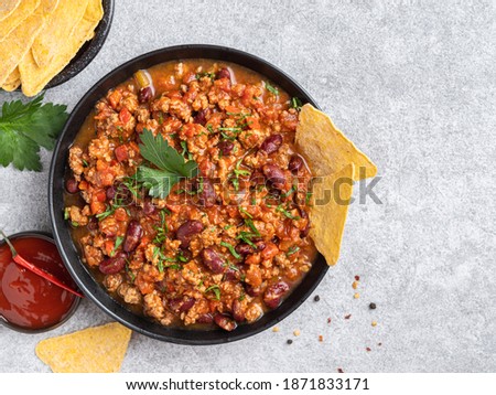 Chilli con carne in black ceramic plate with tortilla corn chips (nachos) and red spicy sauce. Top view, copy space. Grey concrete background. Traditional mexican dish.