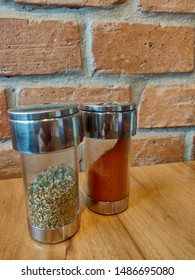 Chilli Pepper And Oregano In Glass Shaker On Wood Table, Brick Background.