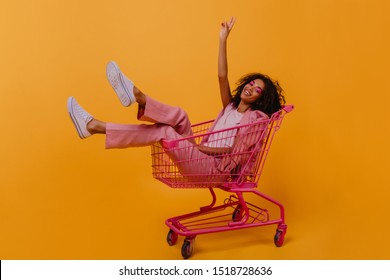 Chilling african girl posing in shopping cart. Indoor photo of emotional young lady in pink clothes.