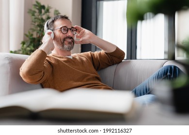 Chilling relaxed mature man father husband listening to the music in headphones lying on the sofa, enjoying sound tracks remotely. Break from working from home.