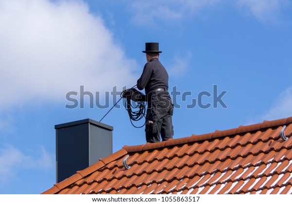 Chimney sweep cleaning a chimney\
standing on the house roof, lowering equipment down the\
flue