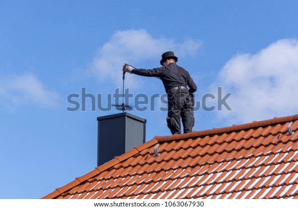 Chimney sweep cleaning a chimney\
standing on the house roof, lowering equipment down the\
flue