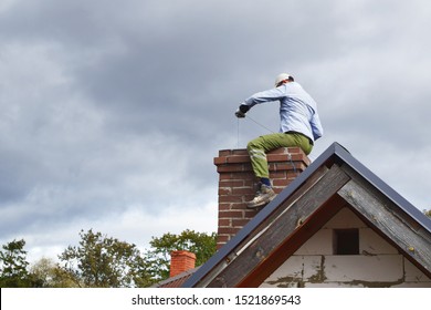 Chimney sweep man cleaning brown brick chimney while sitting on chimney on building roof on cloudy sky background with copy space for text. DIY concept.
