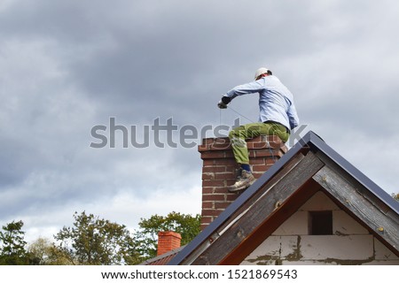 Chimney sweep man cleaning brown brick chimney while sitting on chimney on building roof on cloudy sky background with copy space for text. DIY concept.
