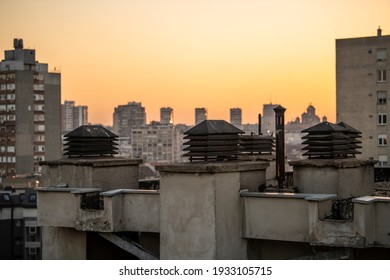 Chimney system on flat rooftop of residential building, with cityscape in background at sunset. Ventilation chimney with a rain cap and shielding. Smokestack pipes.