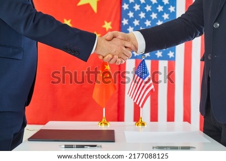 Chinese and American diplomats exchange handshakes. Business representatives from USA and China reach bilateral trade agreement, make deal and shake hands at negotiation table with flags in background