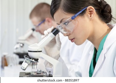 A Chinese Asian female medical or scientific researcher or doctor using a microscope in a laboratory with her colleague out of focus behind her.