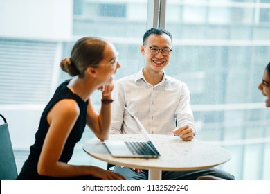 A Chinese Asian man and a Caucasian woman sit at a table and are having a light hearted business discussion during the day in their office. They are part of a diverse team.  - Shutterstock ID 1132529240