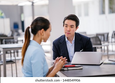 A Chinese Asian manager in a suit has a meeting with his colleague, a woman in a pale blue suit. He is conducting a performance appraisal during this meeting. They are smiling and talking. 