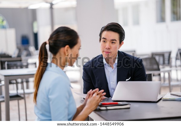 A\
Chinese Asian manager in a suit has a meeting with his colleague, a\
woman in a pale blue suit. He is conducting a performance appraisal\
during this meeting. They are smiling and talking.\
