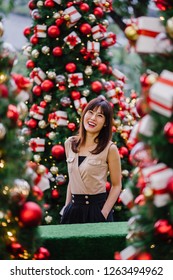 A Chinese Asian tourist woman smiles and poses for a portrait with beautifully decorated Christmas trees during the day in the city. She is slim, elegant and happy as she enjoys the festive mood.