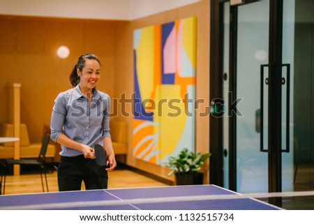 A Chinese Asian woman professional in business attire is playing table tennis (ping pong) in the office with a colleague. She is young and attractive and smiling as she enjoys the break from work. 