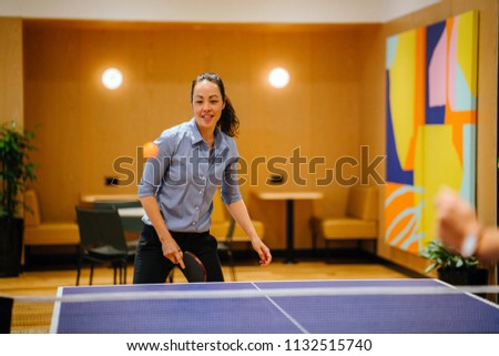 A Chinese Asian woman professional in business attire is playing table tennis (ping pong) in the office with a colleague. She is young and attractive and smiling as she enjoys the break from work. 