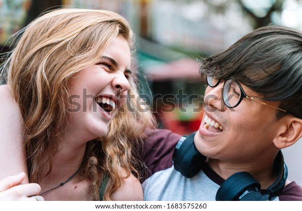 Chinese boy and Caucasian girl\
looking at each other while laughing and hugging on the\
street