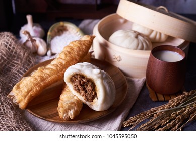 Chinese breakfast: Steamed buns (Baozi) and fried breadsticks (youtiao) on wooden plate, with bamboo steamer in background. soy milk.