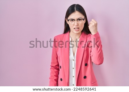 Chinese business young woman wearing glasses angry and mad raising fist frustrated and furious while shouting with anger. rage and aggressive concept. 