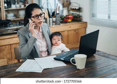 chinese businesswoman sitting in front of computer with infant is arguing with customer on phone. asian female worker holding baby is talking agitatedly on cellphone in her house. - Shutterstock ID 1727705566