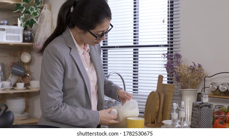 Chinese Career Woman Making Breakfast For Family In Early Morning. Asian Lady In Suit Taking A Jug Of Milk From Fridge And Pouring In Mug While Frying Eggs In Kitchen In Relaxing Mood.