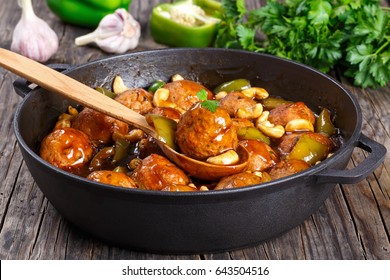 Chinese Cashew Chicken Meatballs Tossed With Sauce Of Green Bell Pepper, Onion, Soy Sauce, Chinese Cooking Wine And Spices In Pan And  One Meatball On Wooden Spoon, Close-up, Ingredients On Table